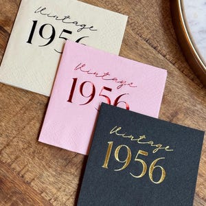 May include: Three square paper napkins with the text "Vintage 1956". One is cream-colored with black text, one is pink with red text, and one is black with gold text. The napkins are on a wooden surface.