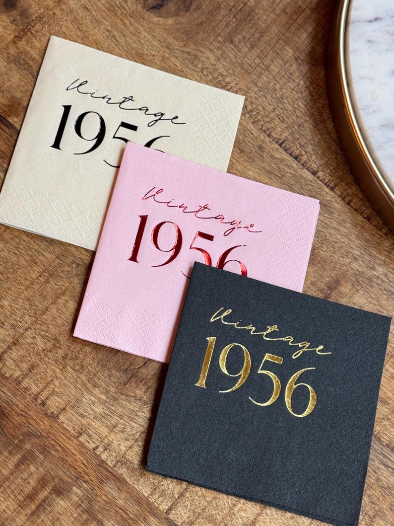May include: Three square paper napkins with the text "Vintage 1956". One is cream-colored with black text, one is pink with red text, and one is black with gold text. The napkins are on a wooden surface.