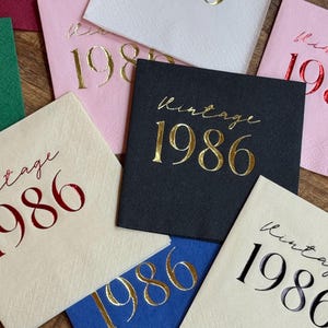 A close-up shows several square paper napkins in various colors, including dark gray, white, pink, blue, and red, all foil-stamped with "Vintage 1986."