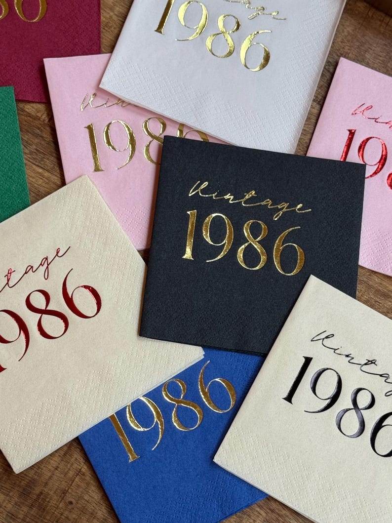 A close-up shows several square paper napkins in various colors, including dark gray, white, pink, blue, and red, all foil-stamped with "Vintage 1986."