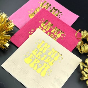May include: Three gold foil napkins with the text "In My Bride Era" in a retro font. The napkins are layered on top of each other, with the top napkin being white, the middle napkin being pink, and the bottom napkin being red.