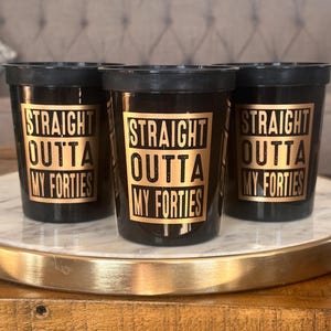 Three black 16oz cups featuring gold bordered text reading Straight Outta My Forties arranged on a tray.