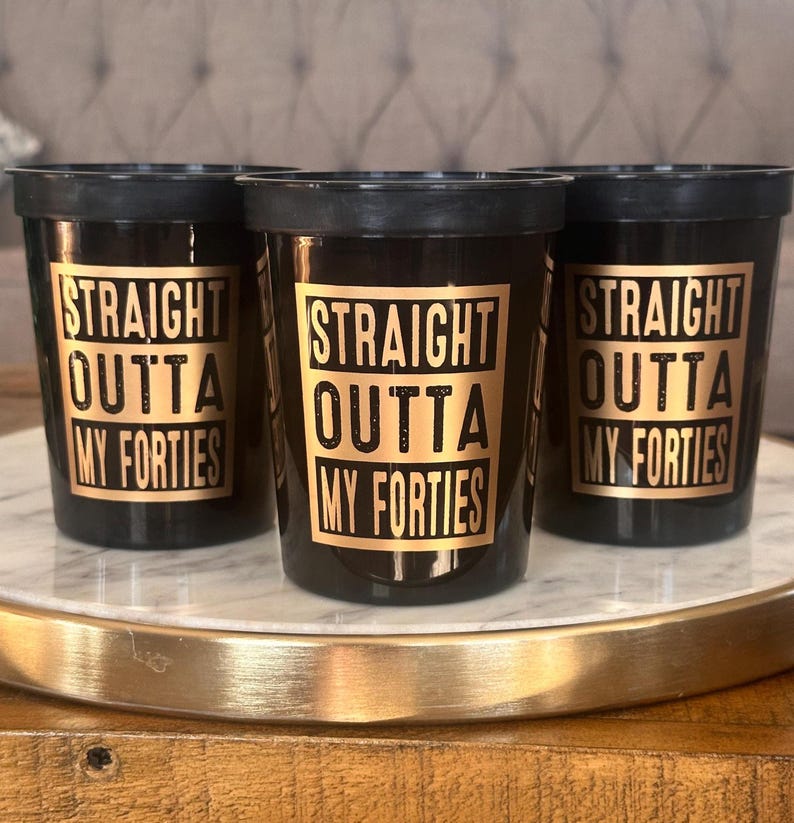 Three black 16oz cups featuring gold bordered text reading Straight Outta My Forties arranged on a tray.