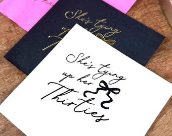 May include: White napkin with black calligraphy that reads "She's tying up her thirties" with a black ribbon bow.