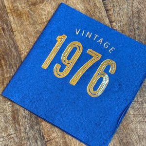 May include: A square, royal blue felt card with the word "VINTAGE" and the number "1976" in gold lettering. The card is displayed on a wooden surface.