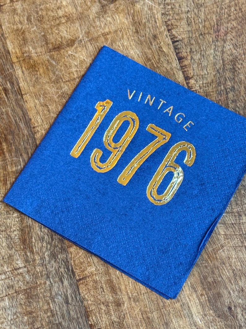 May include: A square, royal blue felt card with the word "VINTAGE" and the number "1976" in gold lettering. The card is displayed on a wooden surface.