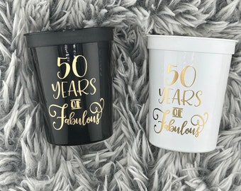 May include: Two plastic cups with black and white exteriors. Both cups have gold lettering that reads "50 Years of Fabulous".