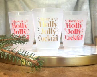 May include: Three frosted plastic cups with the text "Have a Holly Jolly Cocktail" in red and gold lettering. The cups are on a gold-rimmed tray with a sprig of evergreen. Festive drinkware for holiday gatherings.