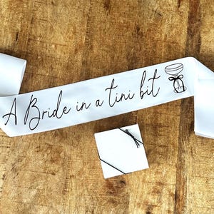 May include: White satin sash with black text that reads "A Bride in a Tini Bit" and a black illustration of a champagne flute with a bow.