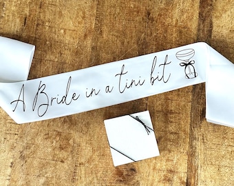 Może przedstawiać: Biała satynowa szarfa z napisem "A Bride in a tini bit" czarnym pismem, wraz z ilustracją kieliszka do martini. Obecne jest również małe białe pudełko na prezent związane czarnym sznurkiem.