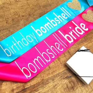 Op de afbeelding: Twee satijnen sjerpen, &eacute;&eacute;n turquoise en &eacute;&eacute;n felroze, met witte tekst. De turquoise sjerp leest "birthday bombshell" en heeft een gouden glitterhart. De roze sjerp leest "bombshell bride" en heeft een gouden glitterhart. Een witte doos met een zwart lint staat in de hoek.