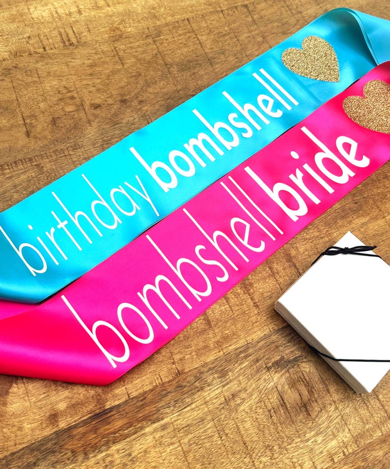 Op de afbeelding: Twee satijnen sjerpen, &eacute;&eacute;n turquoise en &eacute;&eacute;n felroze, met witte tekst. De turquoise sjerp leest "birthday bombshell" en heeft een gouden glitterhart. De roze sjerp leest "bombshell bride" en heeft een gouden glitterhart. Een witte doos met een zwart lint staat in de hoek.