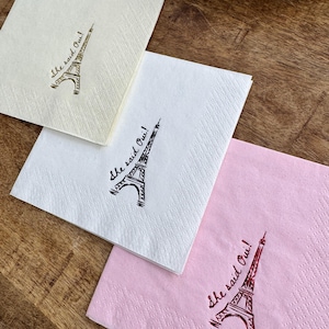 May include: Four square paper napkins in cream, white, and pink, each with a drawing of the Eiffel Tower and the phrase "She said Oui!" in script. The napkins are on a wooden surface.