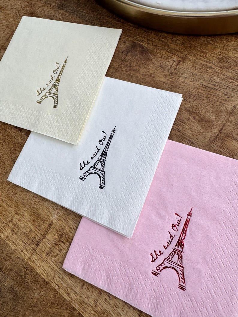 May include: Four square paper napkins in cream, white, and pink, each with a drawing of the Eiffel Tower and the phrase "She said Oui!" in script. The napkins are on a wooden surface.