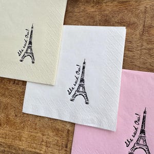 May include: Three square paper napkins in cream, white, and pink, each with a black Eiffel Tower illustration and the phrase "she said Oui!" printed on them. The napkins are arranged on a wooden surface.