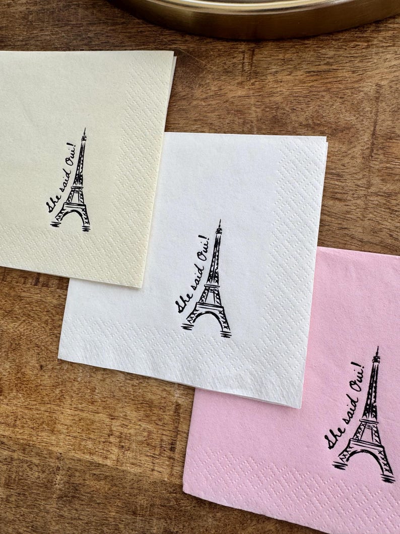 May include: Three square paper napkins in cream, white, and pink, each with a black Eiffel Tower illustration and the phrase "she said Oui!" printed on them. The napkins are arranged on a wooden surface.