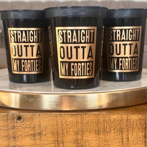 Three black stadium cups with gold text reading Straight Outta My Forties displayed on a decorative tray.