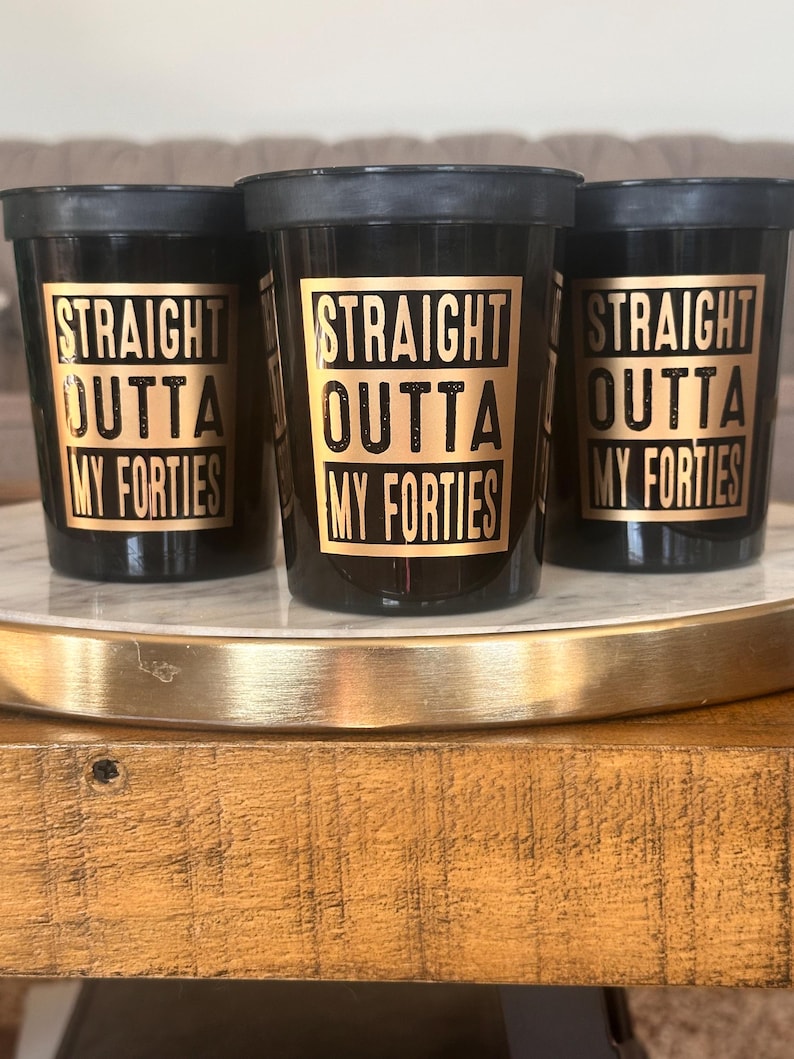 Three black stadium cups with gold text reading Straight Outta My Forties displayed on a decorative tray.