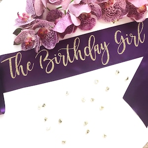 May include: Purple satin sash with gold glitter lettering that reads "The Birthday Girl".