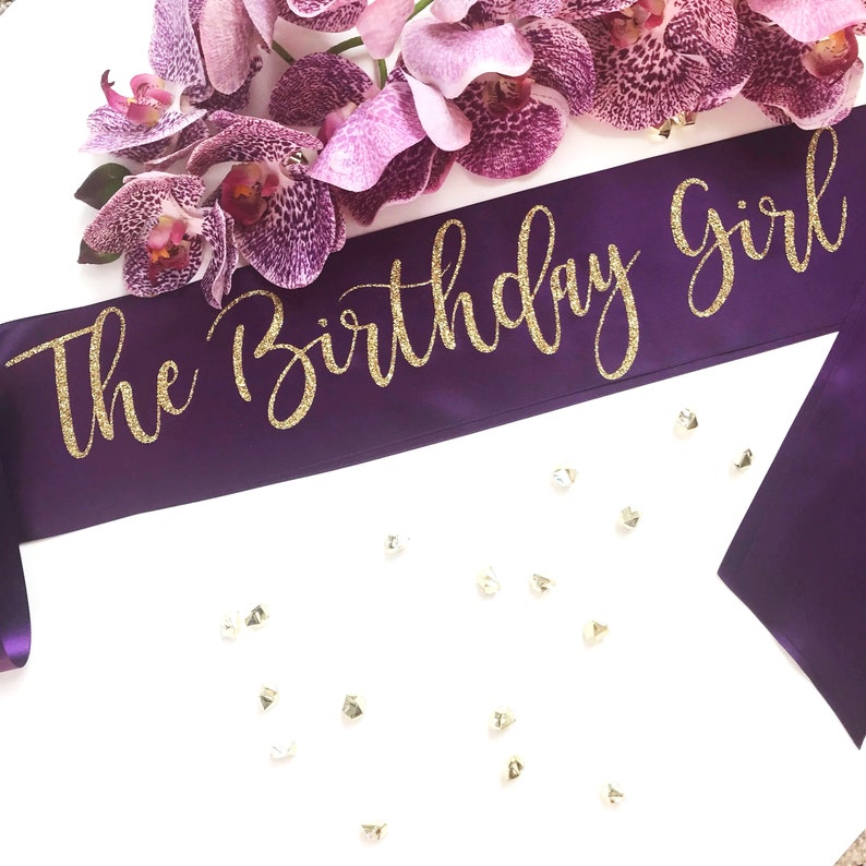 May include: Purple satin sash with gold glitter lettering that reads "The Birthday Girl".