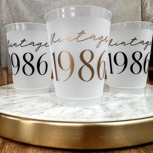 Three frosted plastic cups with "vintage" in script and "1986" in block letters, two with black text and one with gold text, sit on a white marble tray with a gold rim.