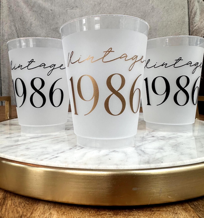 Three frosted plastic cups with "vintage" in script and "1986" in block letters, two with black text and one with gold text, sit on a white marble tray with a gold rim.