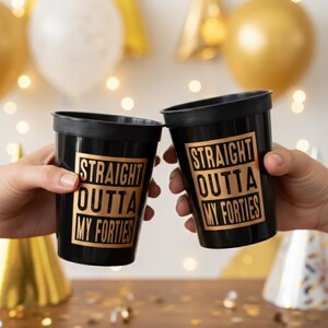 Two hands toast black plastic cups printed with gold text reading Straight Outta My Forties against a party background.