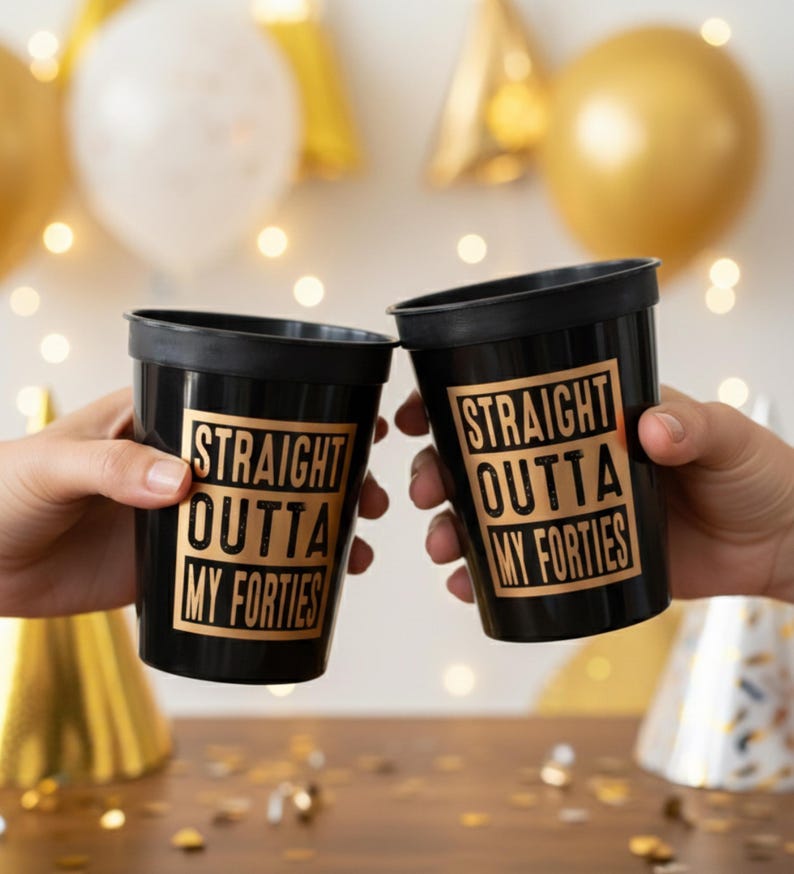 Two hands toast black plastic cups printed with gold text reading Straight Outta My Forties against a party background.