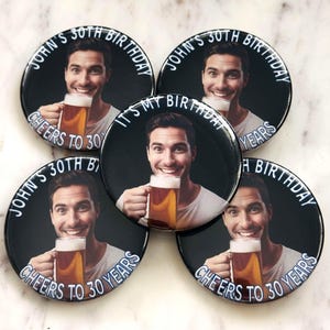 May include: A collection of black round buttons with a photo of a smiling person holding a beer mug. The buttons have white text that reads "John's 30th Birthday" and "Cheers to 30 Years."