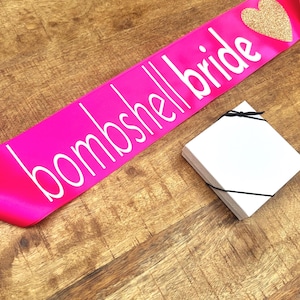Op de afbeelding: Een felroze satijnen sjerp met de tekst "bombshell bride" in witte letters en een gouden glitterhart. Een stapel witte kaarten ligt naast de sjerp. De items liggen op een houten oppervlak.