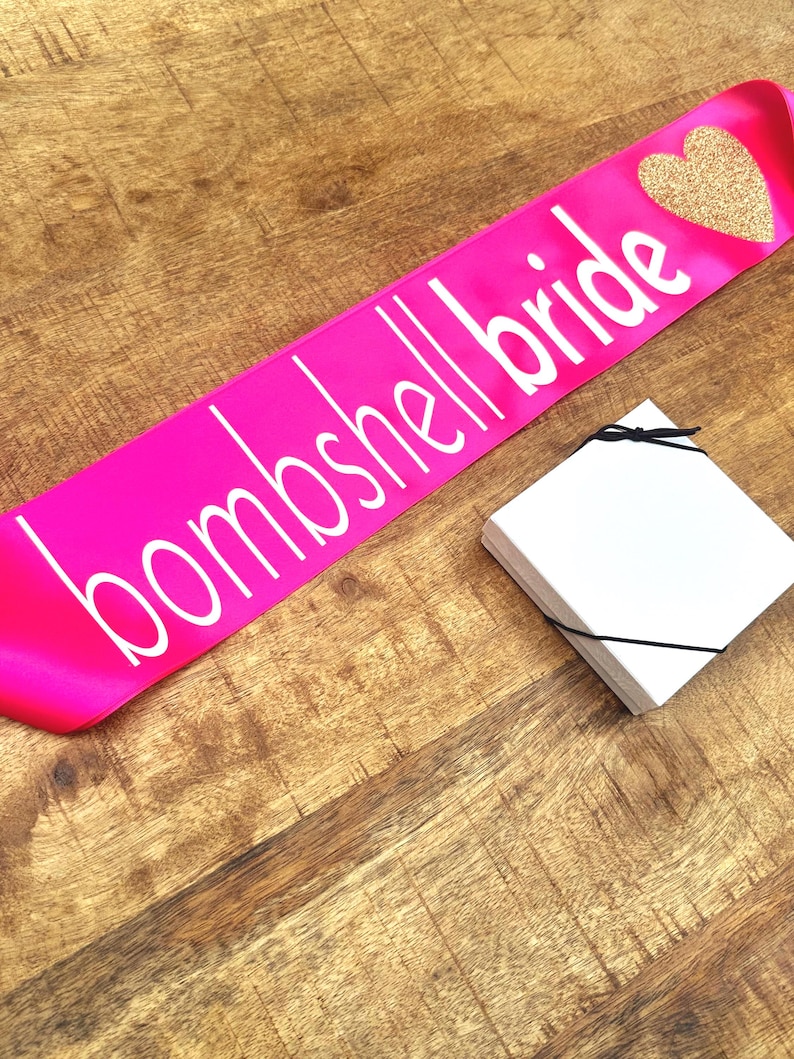 Op de afbeelding: Een felroze satijnen sjerp met de tekst "bombshell bride" in witte letters en een gouden glitterhart. Een stapel witte kaarten ligt naast de sjerp. De items liggen op een houten oppervlak.