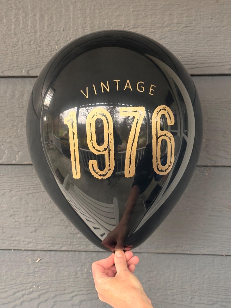 Puede incluir: Un globo negro con el texto dorado "VINTAGE 1976". El globo refleja el entorno y est&aacute; sostenido por una mano. Es una decoraci&oacute;n para fiestas.
