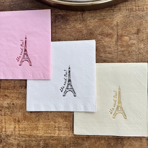 May include: Four square paper napkins in pink, white, and cream, each with a drawing of the Eiffel Tower and the phrase "She said Oui!" in a matching colour. The napkins are arranged on a wooden surface.