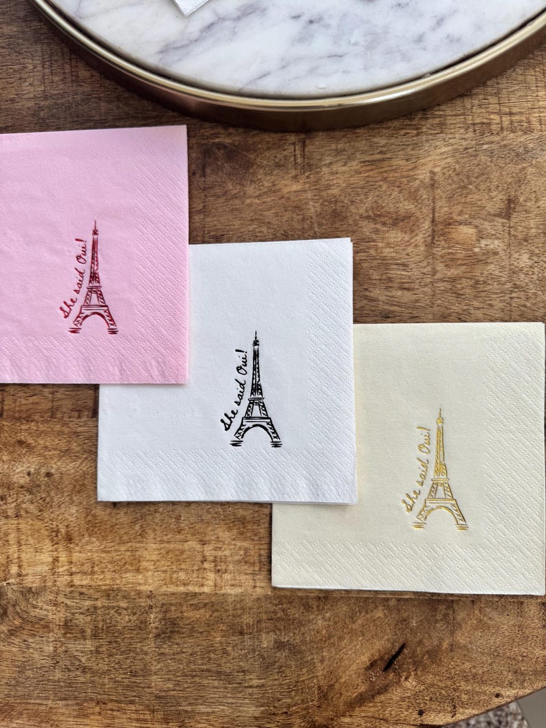 May include: Four square paper napkins in pink, white, and cream, each with a drawing of the Eiffel Tower and the phrase "She said Oui!" in a matching colour. The napkins are arranged on a wooden surface.