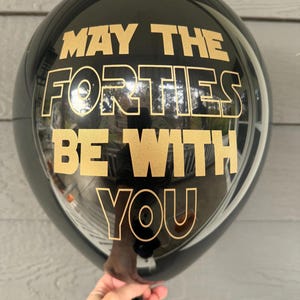 A person holding a round black balloon printed with gold text that reads May the Forties be with you.