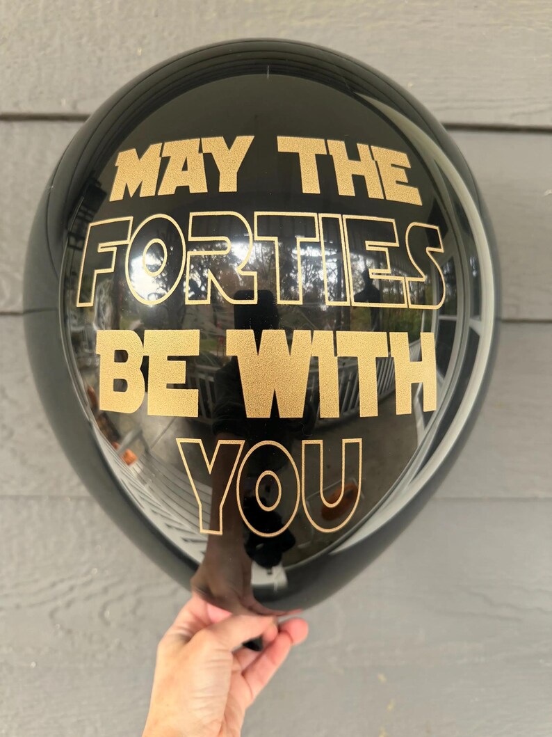 A person holding a round black balloon printed with gold text that reads May the Forties be with you.