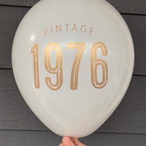 Puede incluir: Un globo blanco con la palabra "VINTAGE" encima del n&uacute;mero dorado "1976". El globo est&aacute; sostenido por una mano y est&aacute; contra un fondo gris oscuro. Este globo es perfecto para una celebraci&oacute;n de cumplea&ntilde;os.