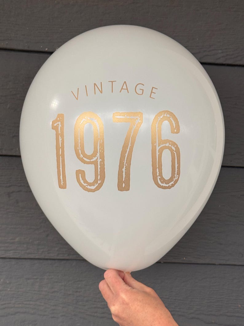 Puede incluir: Un globo blanco con la palabra "VINTAGE" encima del n&uacute;mero dorado "1976". El globo est&aacute; sostenido por una mano y est&aacute; contra un fondo gris oscuro. Este globo es perfecto para una celebraci&oacute;n de cumplea&ntilde;os.