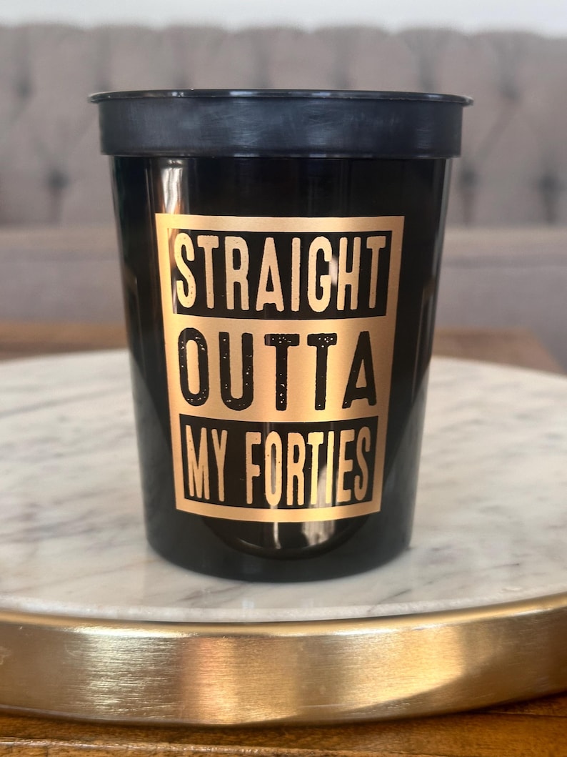 Close-up of one black plastic party cup with gold metallic text reading Straight Outta My Forties.