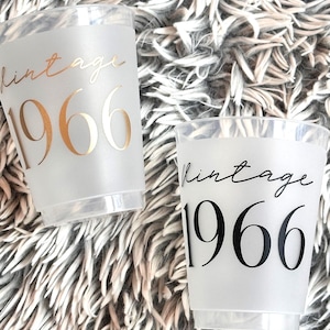 May include: Two clear plastic cups with a frosted finish. The cups are printed with the word "vintage" and the number "1966" in gold and black lettering. The cups are on a fluffy, textured surface.