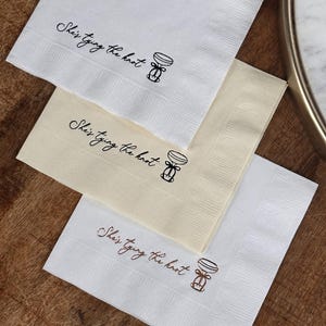 Bachelorette party cocktail napkins with the text "She's tying the knot" and a simple illustration of a champagne flute with a bow. The napkins are stacked on top of each other.