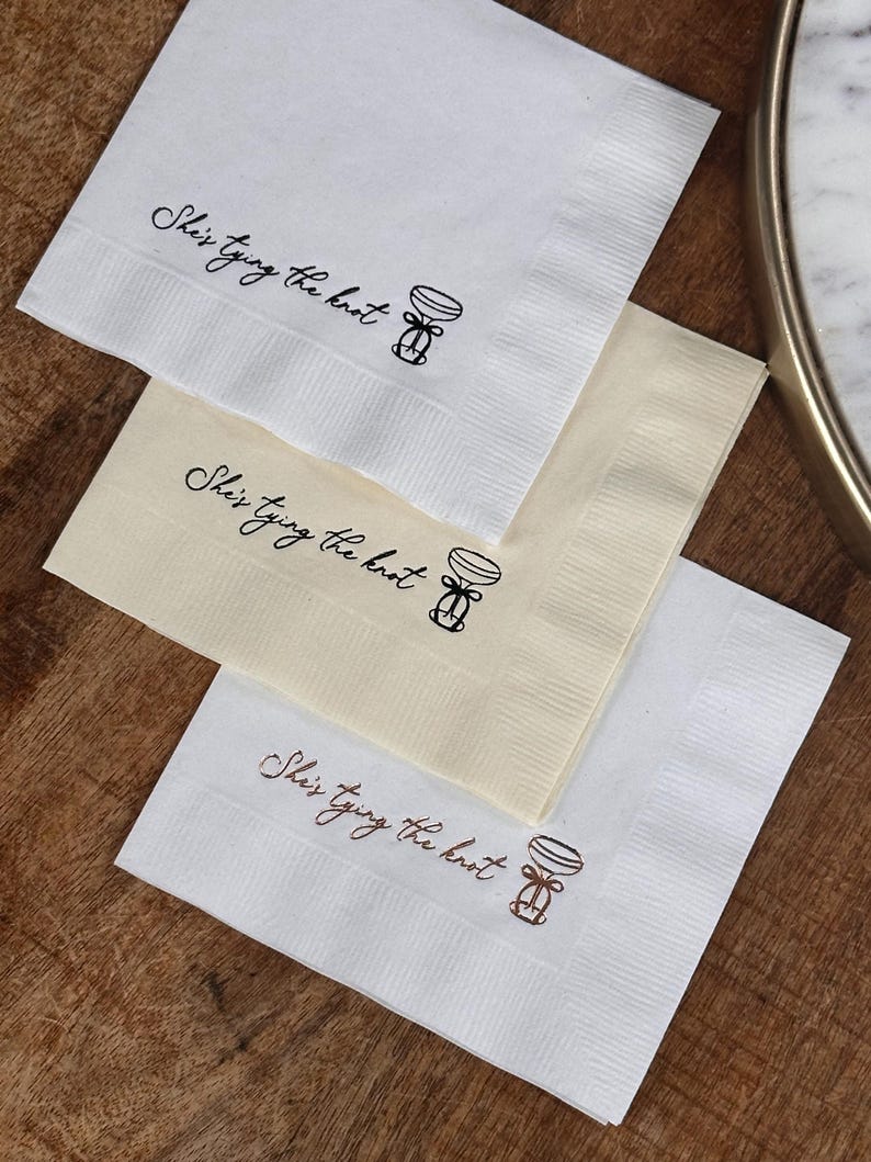 Bachelorette party cocktail napkins with the text "She's tying the knot" and a simple illustration of a champagne flute with a bow. The napkins are stacked on top of each other.