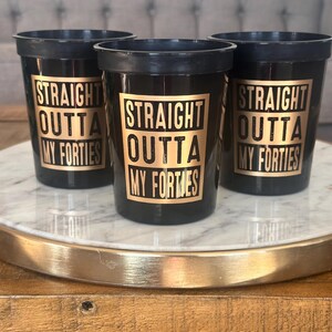 A trio of black plastic cups displaying gold typography reading Straight Outta My Forties on a circular tray.