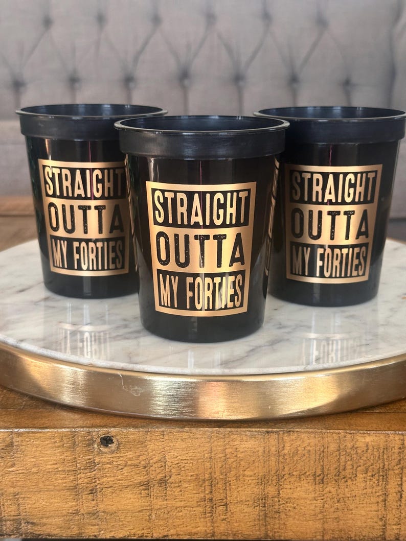 A trio of black plastic cups displaying gold typography reading Straight Outta My Forties on a circular tray.