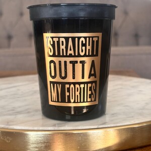 A black plastic cup with gold lettering reading Straight Outta My Forties positioned on a white marble surface.