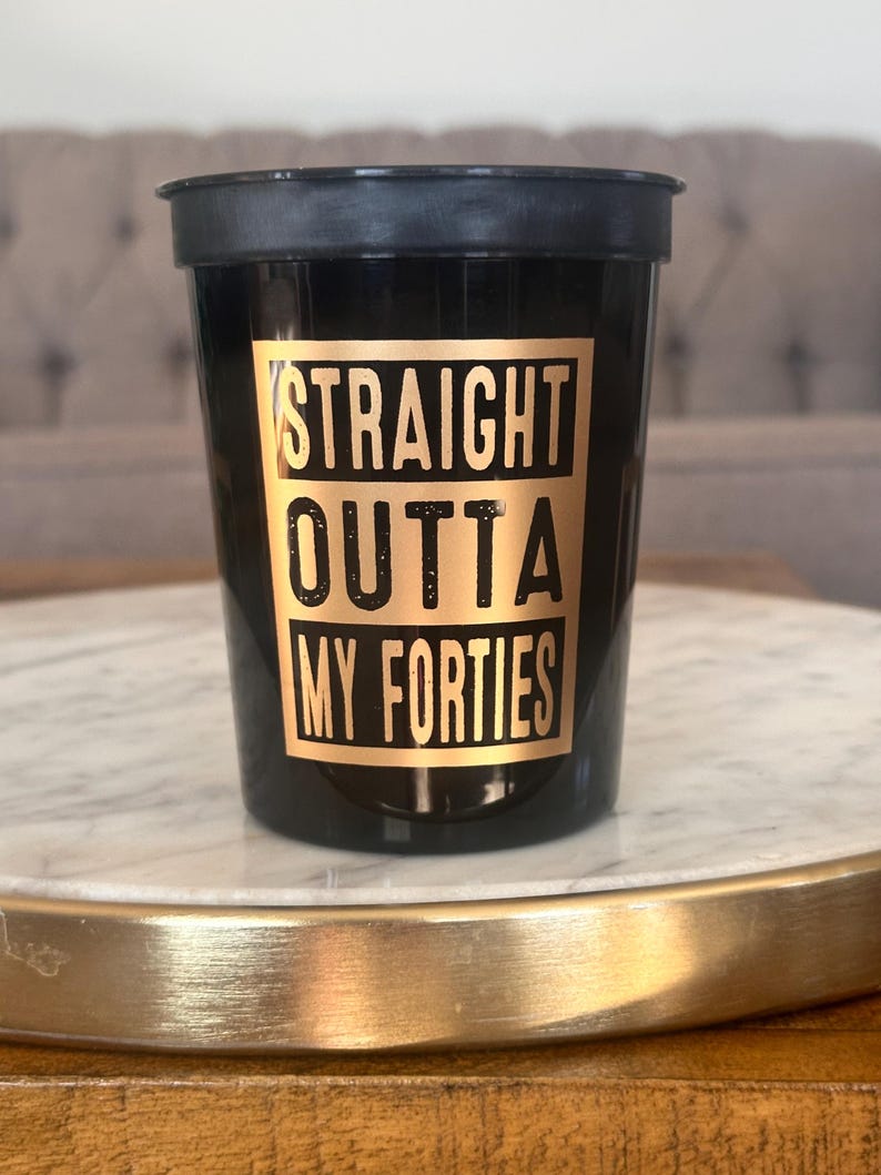 A black plastic cup with gold lettering reading Straight Outta My Forties positioned on a white marble surface.