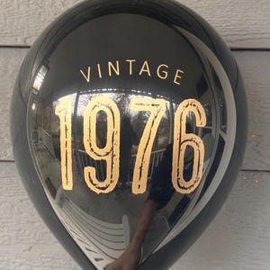 Puede incluir: Un globo negro con el texto dorado "VINTAGE 1976". El globo es redondo y brillante, reflejando el entorno. El texto est&aacute; en una fuente serif.
