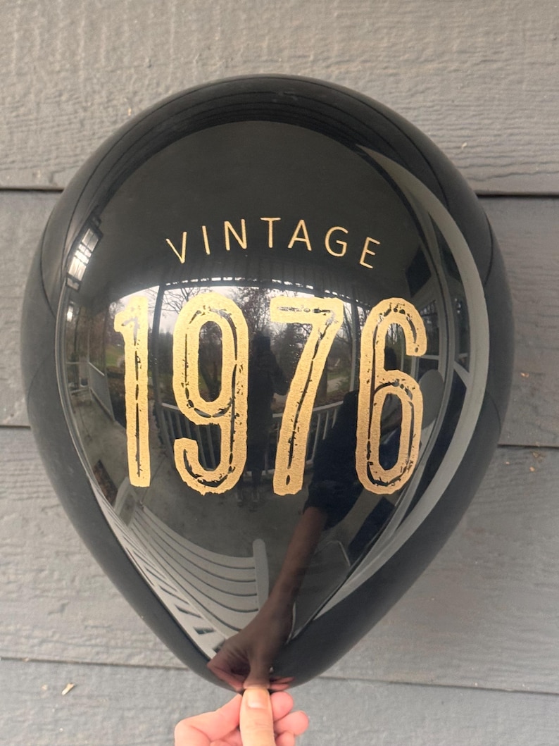 Puede incluir: Un globo negro con el texto dorado "VINTAGE 1976". El globo es redondo y brillante, reflejando el entorno. El texto est&aacute; en una fuente serif.