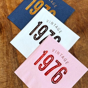 May include: Three square napkins with the word "VINTAGE" above the year. The top napkin is navy blue with gold lettering for "1970". The middle napkin is white with brown lettering for "1970". The bottom napkin is pink with red lettering for "1976".