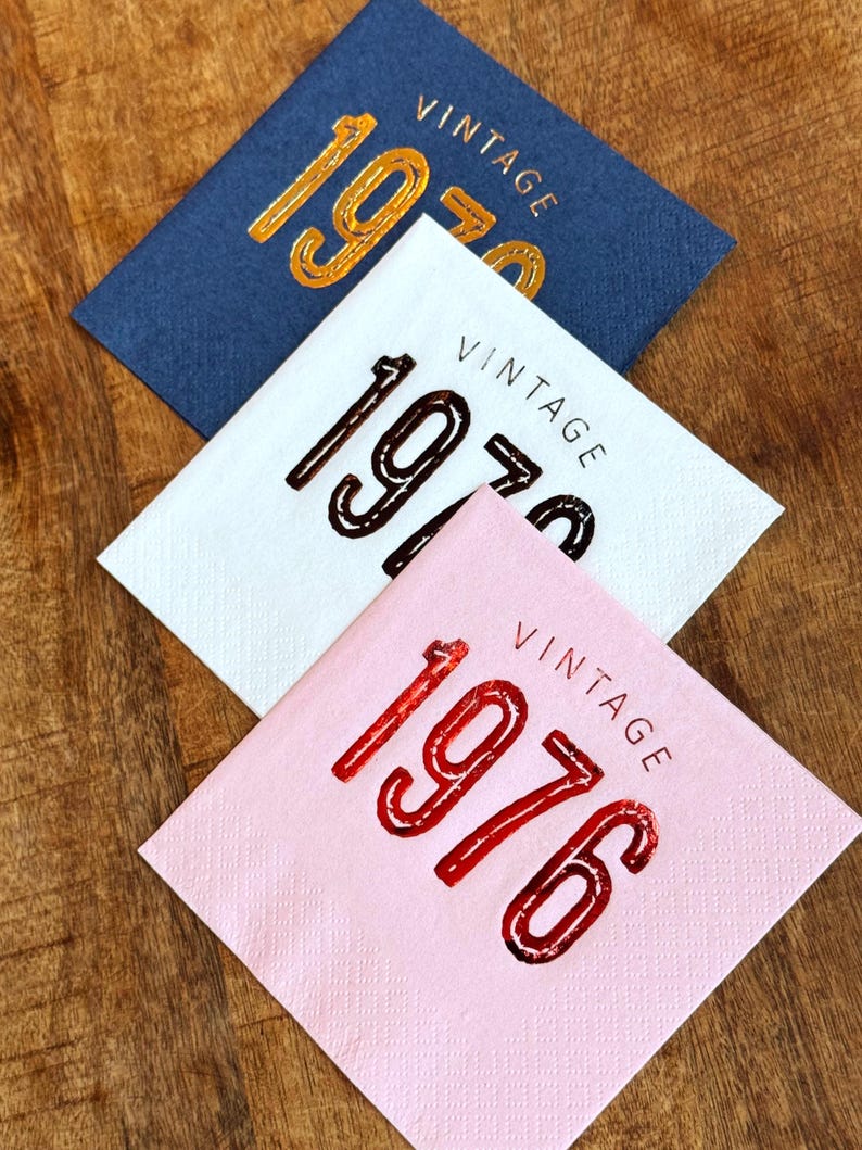 May include: Three square napkins with the word "VINTAGE" above the year. The top napkin is navy blue with gold lettering for "1970". The middle napkin is white with brown lettering for "1970". The bottom napkin is pink with red lettering for "1976".